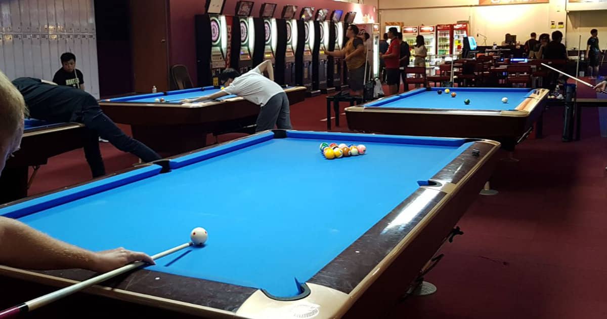 Players at blue-cloth pool tables in a busy pool hall, with electronic dartboards and drinks fridges along the walls